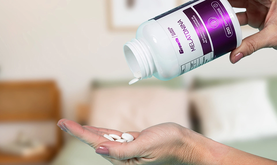 Uma mão esquerda de uma mulher com comprimidos na mão, e na mão direita a embalagem da melatonina 0,21mg (200 Comprimidos) - Growth Supplements