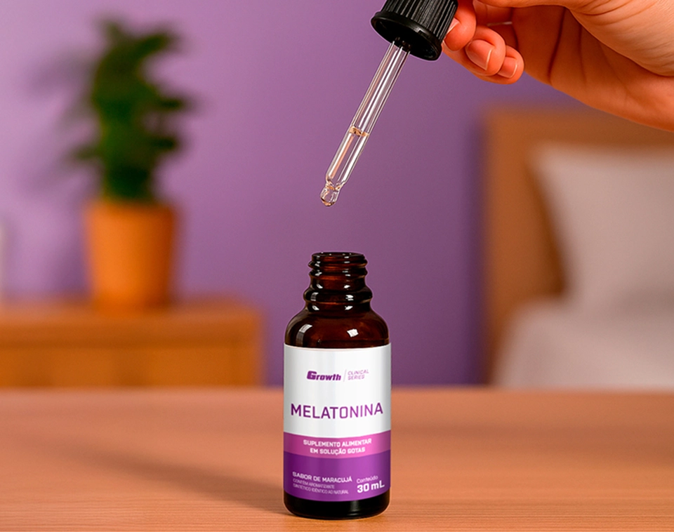 Uma mão esquerda de uma pessoa com o conta-gotas na mão, e embaixo a Melatonina 0,21mg 30ml sabor maracujá - growth supplements em cima de uma mesa