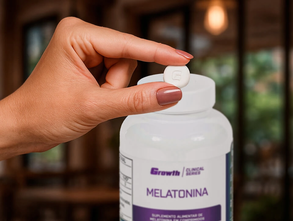 Uma mão de uma mulher segurando um comprimido e no fundo a embalagem da melatonina 0,21mg (100 Comprimidos) - Growth Supplements
